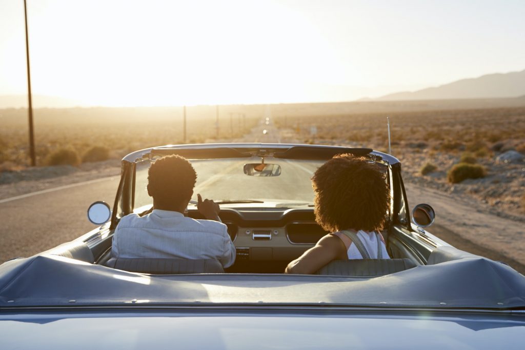 Rear View Of Couple On Road Trip Driving Classic Convertible Car Towards Sunset